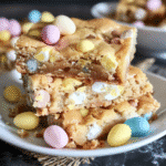 Cadbury Mini Egg Blondie Recipe 11 Stacked Cadbury Mini Egg Blondie Recipe on a plate, surrounded by pastel chocolate eggs
